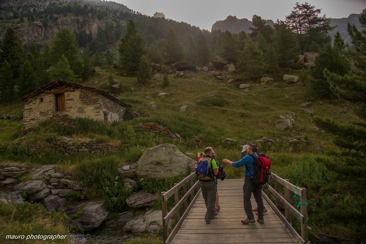 Escursionisti su un ponte di legno che supera il torrente vallanta; sulla sinistra una baita in pietra, sullo sfondo prato verde e larici