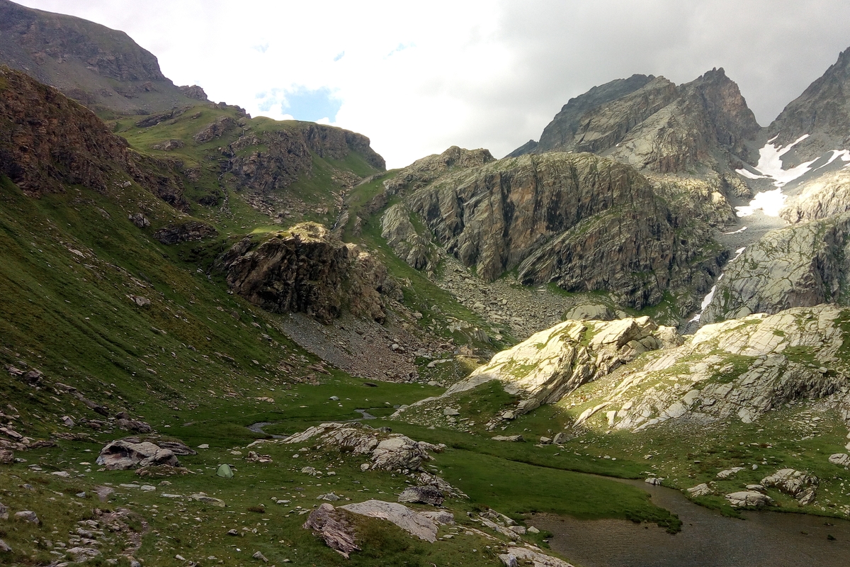 ambiente montano di alta quota con a sinistra prateria alpina e versanti inerbiti, a destra rocce e torrente che sfocia in un lago