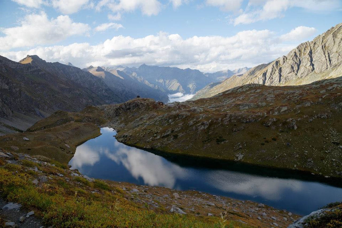 Un lago alpino dalla forma allungata in cui si specchiano le nuvole, intorno imponenti montagne, cielo azzurro.